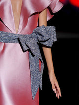 Vestido de fiesta largo en mikado pique rosa loto, con escote en V, volantes en ambos lados y falda sirena entallada, cinturon con lazo azul noche de HANNIBAL LAGUNA.