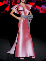 Vestido de fiesta largo en mikado pique rosa loto, con escote en V, volantes en ambos lados y falda sirena entallada, cinturon con lazo azul noche de HANNIBAL LAGUNA.