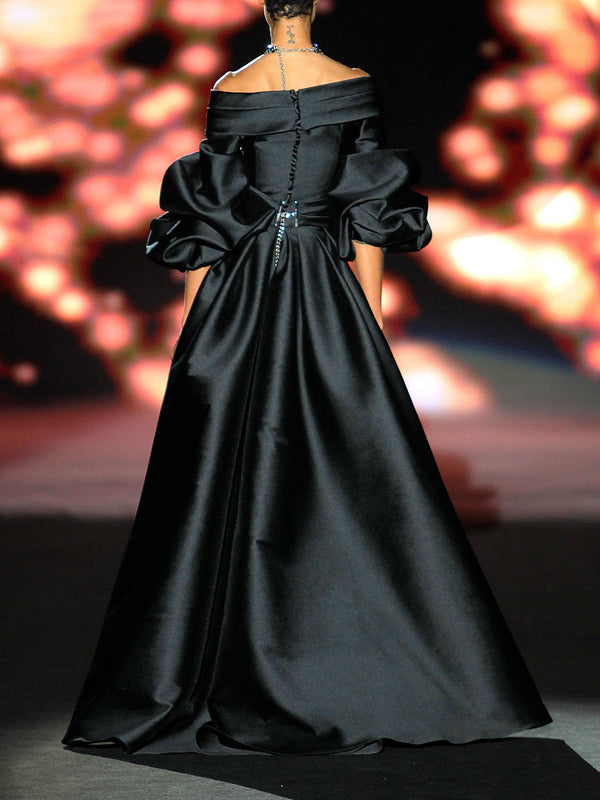 Vestido de fiesta realizado en matelé negro con escote liz cruzado, maxi mangas abullonadas y falda compuesta de sirena con pliegues y amplia cola de Hannibal Laguna